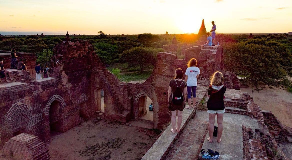 Un viaje en grupo de WeRoad contempla el atardecer desde la cima de una antigua ruina de templo de ladrillo, con vistas a un paisaje verde.