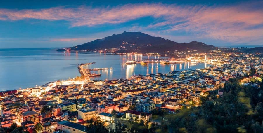 Una vista panoramica di una città costiera illuminata e del suo porto al tramonto, con una grande montagna stagliata contro un cielo colorato.
