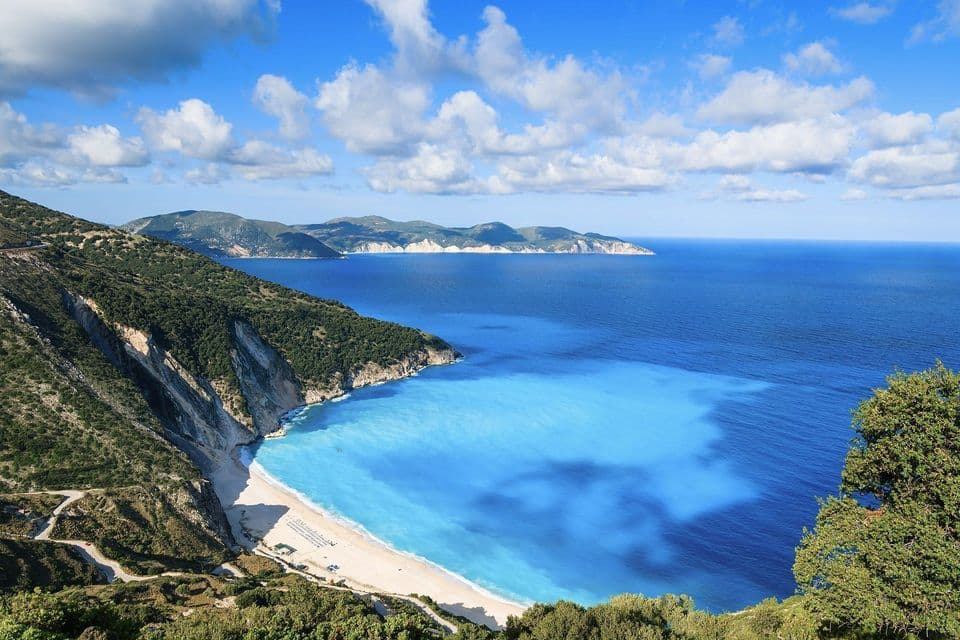 Vista dall'alto di una spiaggia di sabbia bianca e di un mare turchese in una cala, incorniciata da ripide scogliere verdi sotto un cielo parzialmente nuvoloso.