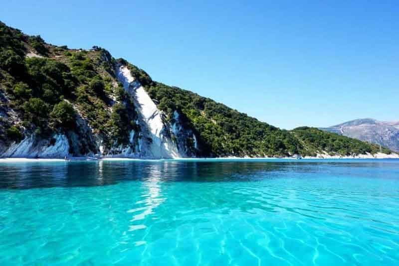 Acqua cristallina e turchese si infrange su una spiaggia bianca ai piedi di una scogliera verde e ripida sotto un cielo blu senza nuvole.