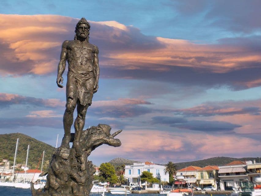Un'alta statua di bronzo di un guerriero si erge in un porto, con una città costiera e colline sullo sfondo sotto un cielo nuvoloso al tramonto.