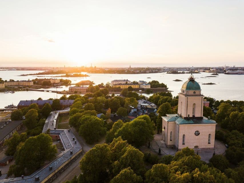 Vue aérienne d'une église au dôme vert sur une île boisée, avec le soleil couchant sur l'eau et une ligne d'horizon urbaine au loin.