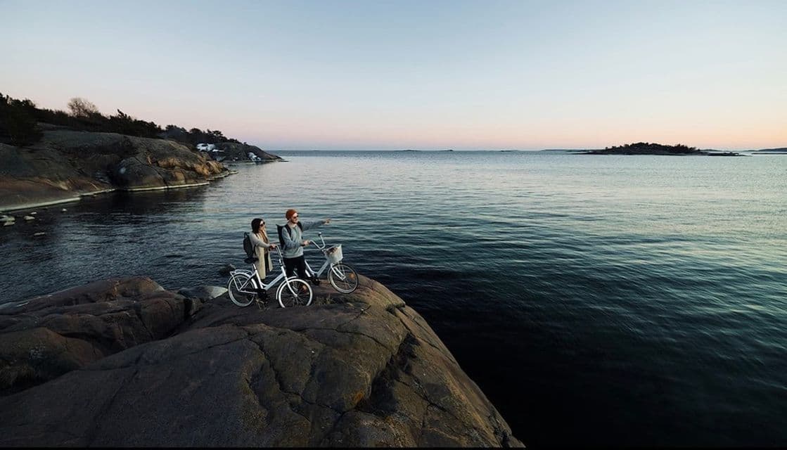 Un couple à vélo se tient sur une falaise rocheuse dominant la mer, l'homme pointant du doigt l'horizon au coucher du soleil.