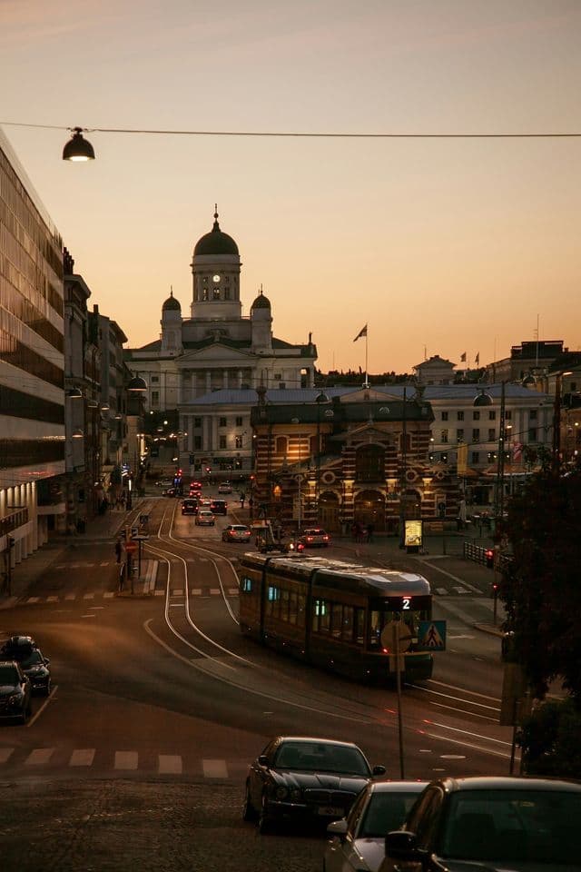 Un tramway circule sur des rails dans une rue de ville au coucher du soleil, avec une grande cathédrale blanche visible en arrière-plan.