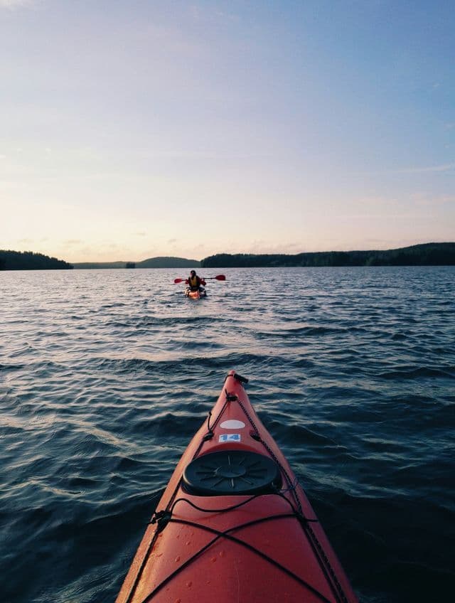 Vue à la première personne depuis un kayak rouge lors d'un voyage de groupe WeRoad, traversant un lac en direction d'un autre kayakiste au crépuscule.