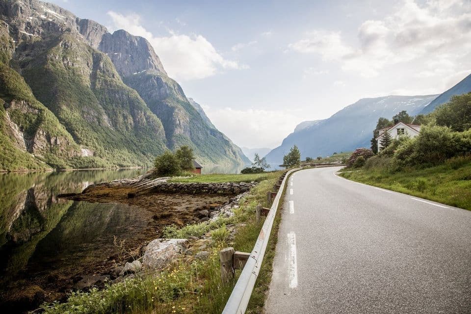 Eine asphaltierte Straße schlängelt sich am Ufer eines Fjords entlang, aus dem steile, grüne Berge unter einem teils bewölkten Himmel emporragen.