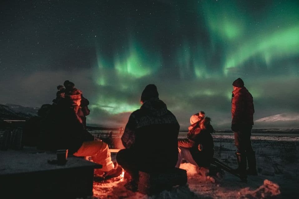 Un grupo de WeRoad se sienta alrededor de una hoguera en un paisaje nevado de noche, observando la aurora boreal verde en el cielo estrellado.