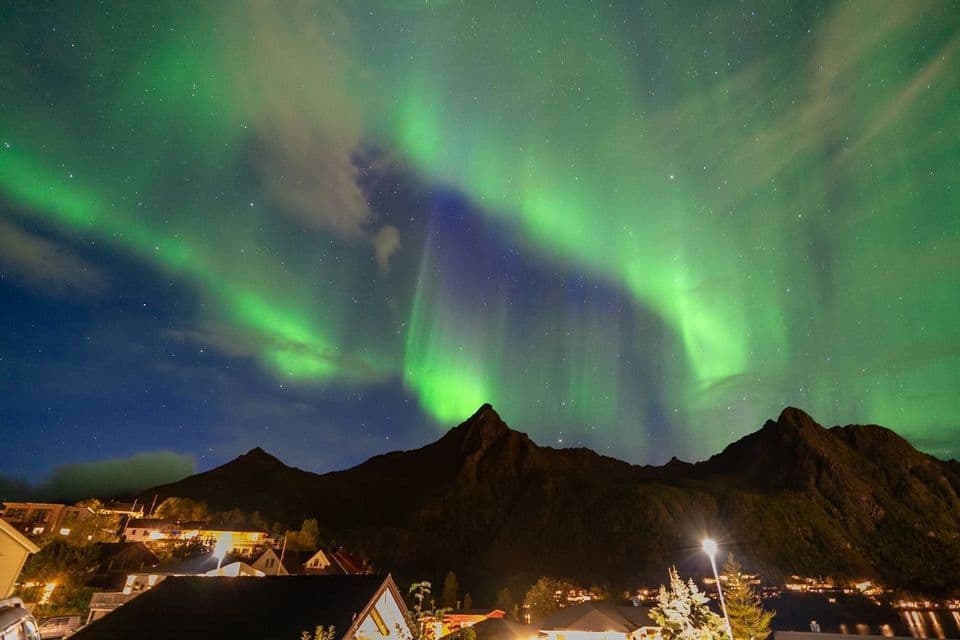 Les aurores boréales vertes et violettes brillent dans un ciel nocturne étoilé, au-dessus de la silhouette sombre de montagnes et d'un village illuminé.