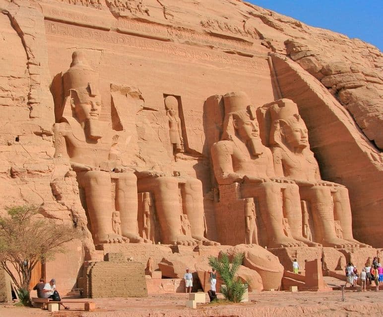 Kolossale antike Statuen, die in eine Sandsteinfelswand gehauen sind, mit Touristen, die an einem sonnigen Tag am Fuß spazieren gehen.
