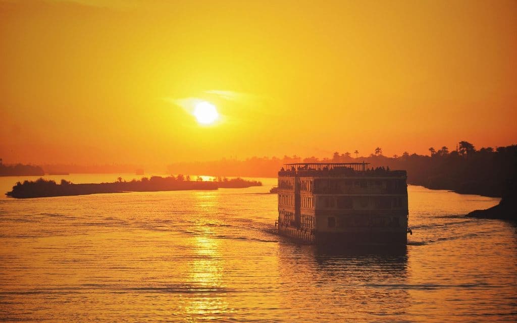 Ein Flussschiff fährt auf einem ruhigen Fluss, beleuchtet von einem goldenen Sonnenuntergang.