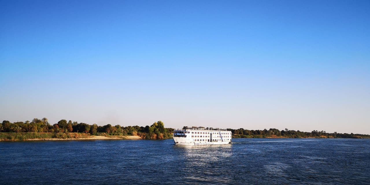 Ein weißes Kreuzfahrtschiff fährt auf einem breiten Fluss mit baumbestandenen Ufern unter einem klaren blauen Himmel.