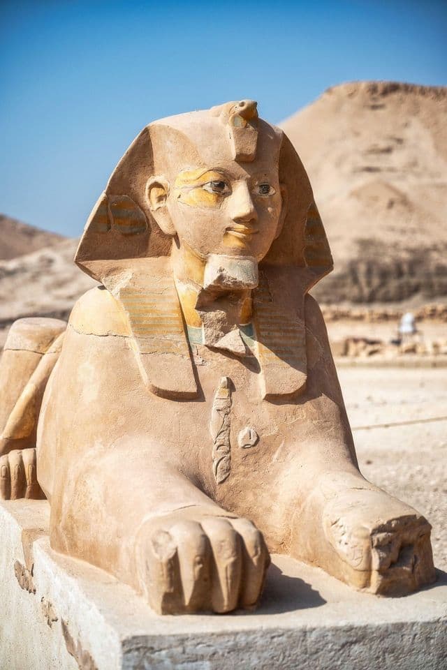 Eine antike Sandstein-Sphinx-Statue mit traditioneller Kopfbedeckung steht auf einem Steinsockel in einer sonnigen Wüstenlandschaft.