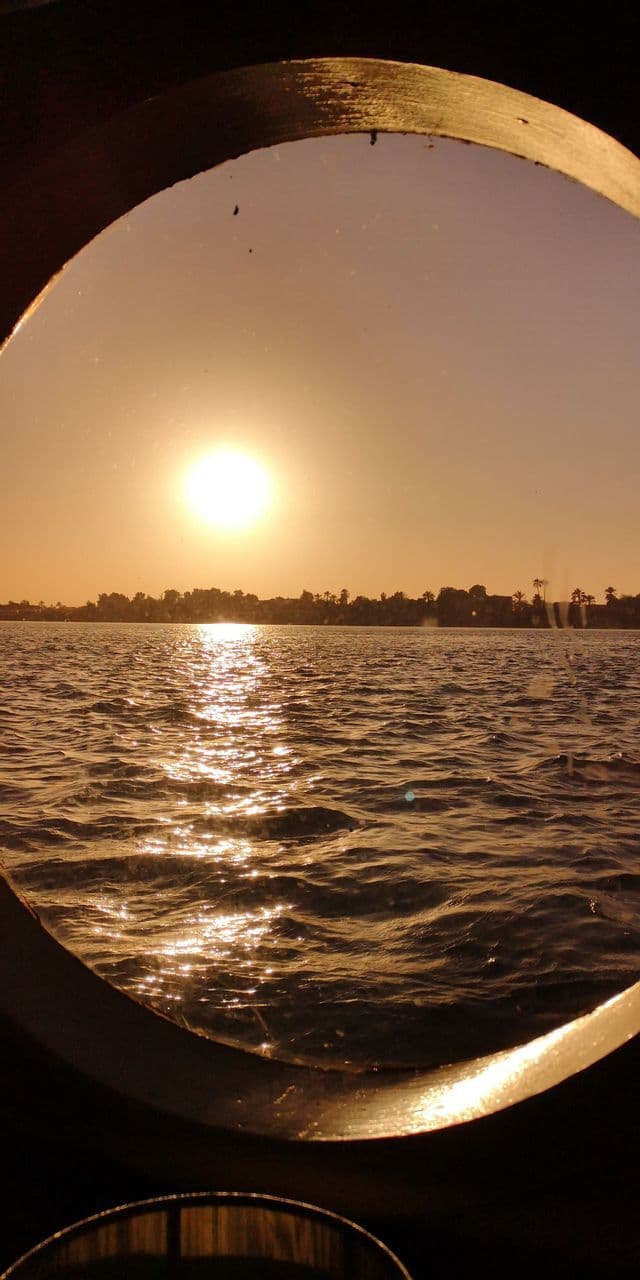 Eine helle Sonne, die über dem Wasser mit fernen Bäumen untergeht, gesehen durch ein rundes Schiffsfenster.