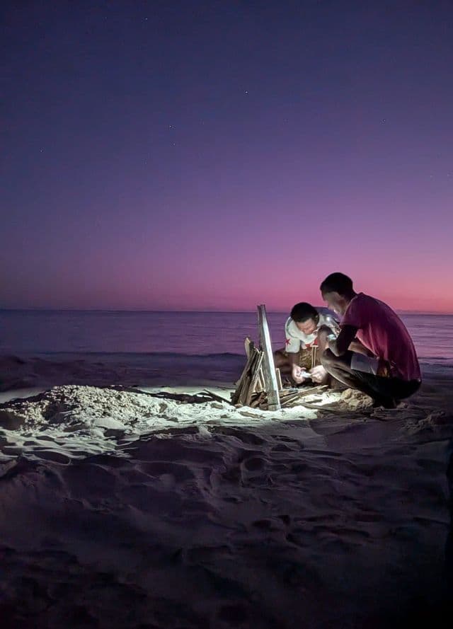 Deux personnes accroupies sur une plage de sable au crépuscule, préparent un petit feu de joie avec du bois flotté au bord de l'océan sous un ciel pourpre.