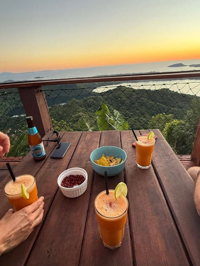 Un voyage de groupe WeRoad savourant des boissons et des en-cas sur une table en bois, avec vue sur un paysage tropical et la mer au coucher du soleil.