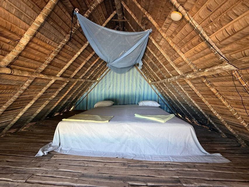 Un lit avec une moustiquaire suspendue dans une chambre mansardée rustique avec un toit de chaume et un plancher en bois.