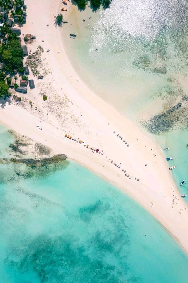 Vue aérienne d'un banc de sable blanc s'étendant dans une eau turquoise limpide, avec des bateaux et un petit village côtier visibles.