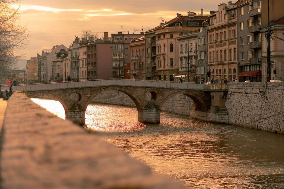 Un ponte ad arco in pietra attraversa un fiume che scorre attraverso una città, con la luce dorata del tramonto che si riflette sull'acqua.