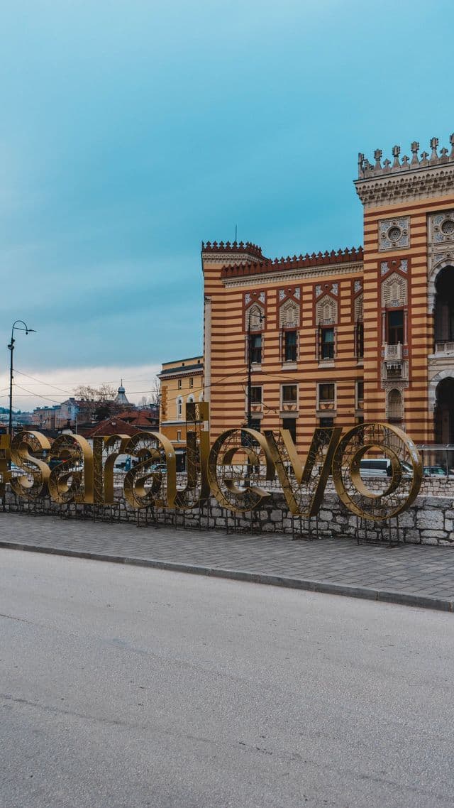 Una grande insegna dorata 'Sarajevo' si erge su un muro di pietra davanti a un edificio a strisce ornato sotto un cielo nuvoloso.