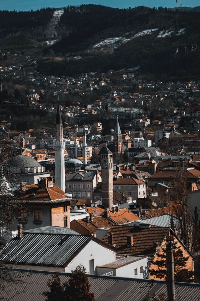 Un paesaggio urbano denso con tetti di tegole rosse, un minareto e una torre dell'orologio, incastonato contro una montagna boscosa con chiazze di neve.