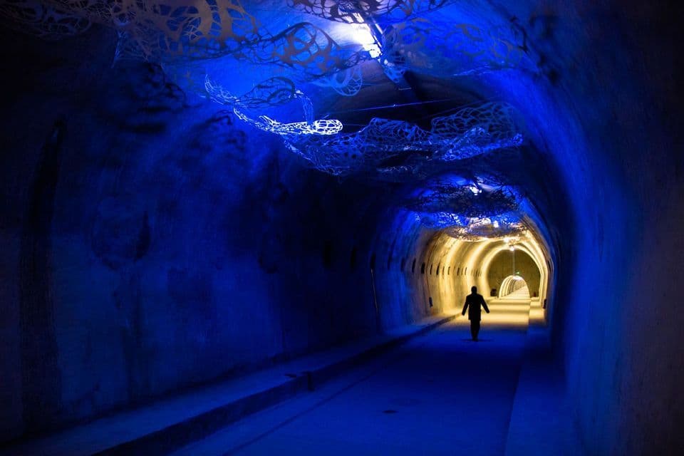 A person walks through a long tunnel illuminated in blue, with a white abstract art installation hanging from the ceiling.