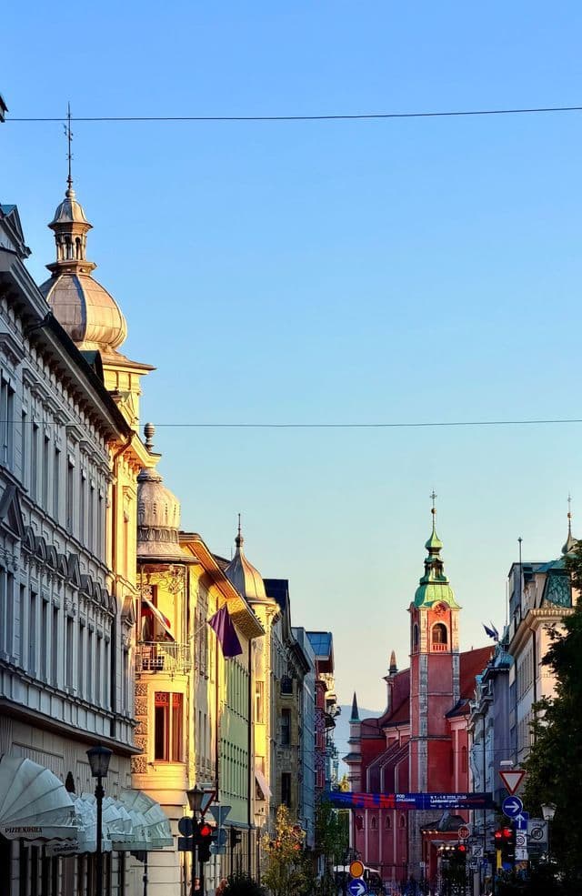 Ornate city buildings and a distant church steeple are lit by warm sunlight against a clear blue sky.