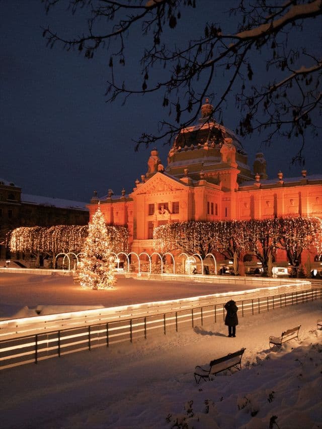 A large, ornate building and a Christmas tree are brightly lit at night above a snowy square with an ice rink.