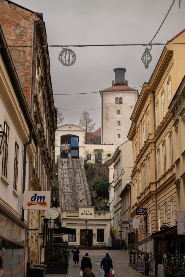 A view up a narrow cobblestone street towards a funicular ascending a hill between old buildings, with a tower visible in the background.