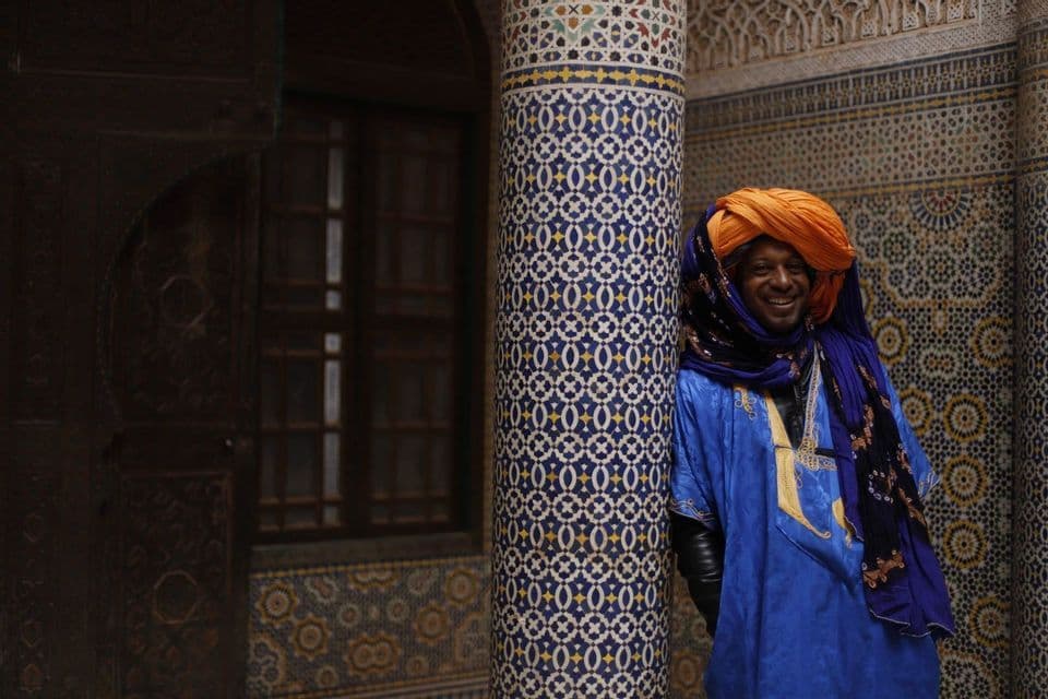 A smiling man wearing a blue robe and an orange turban leans against a pillar covered in intricate blue and yellow mosaic tiles.