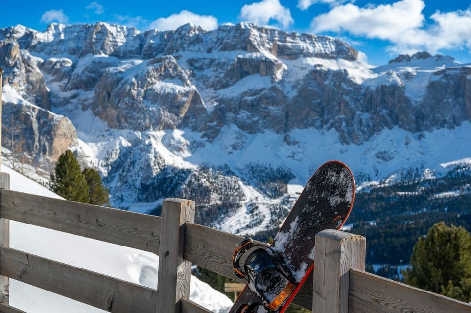Uno snowboard è appoggiato a una staccionata di legno, con una vista panoramica su una catena montuosa innevata sotto un cielo blu.