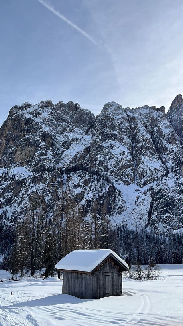 Una piccola baita di legno con il tetto coperto di neve si trova in un paesaggio innevato ai piedi di un'imponente montagna rocciosa.