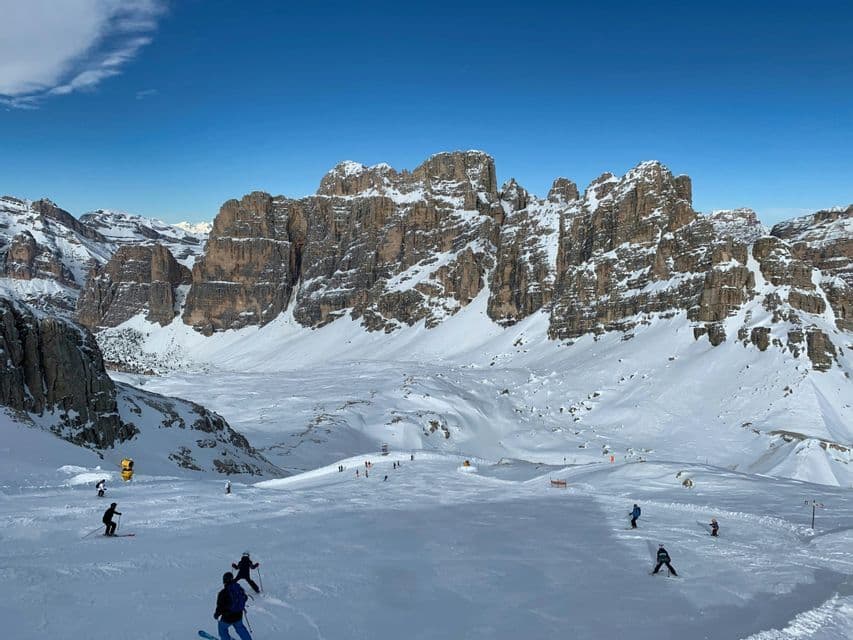Diverse persone sciano su un'ampia pista innevata ai piedi di una catena montuosa frastagliata in una giornata di sole.