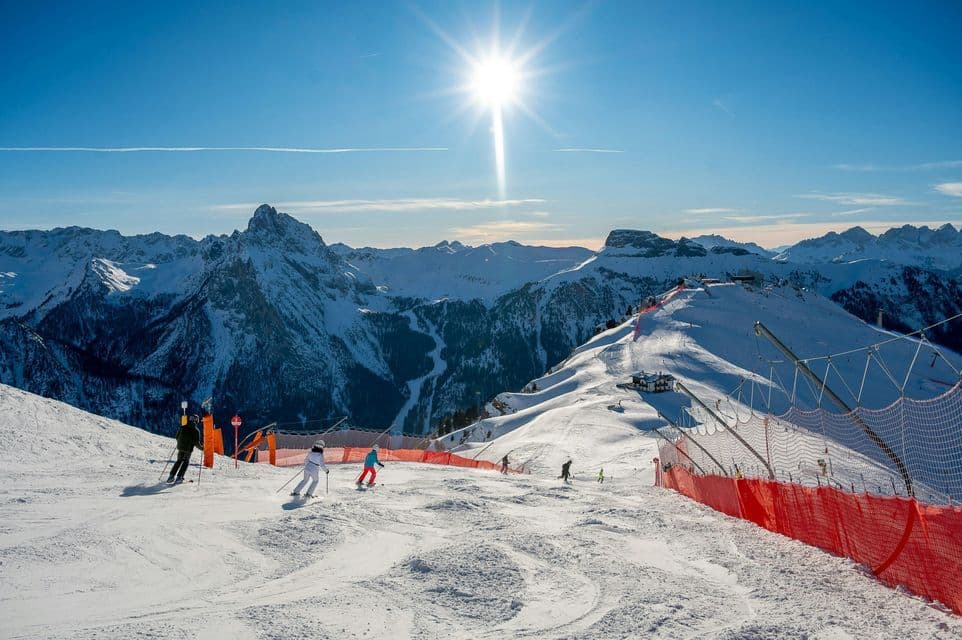 Sciatori scivolano lungo un'ampia pista innevata tra le montagne, sotto un sole splendente e un cielo azzurro limpido.