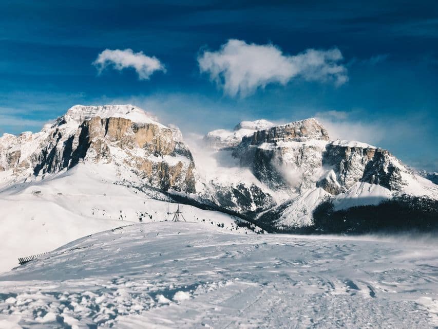Una vasta catena montuosa innevata sotto un cielo azzurro brillante, con il vento che soffia la neve dalle vette.