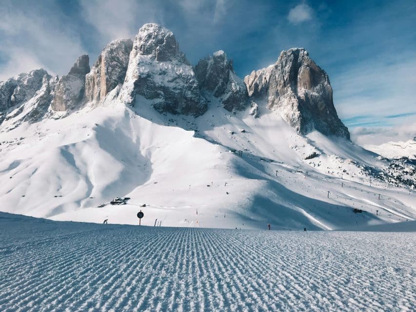 Una pista da sci appena battuta con le sue caratteristiche righe a velluto, che conduce verso una catena di montagne frastagliate e innevate sotto un cielo azzurro.