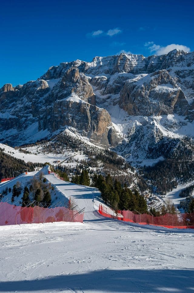 Una pista da sci battuta con reti di sicurezza rosse scende verso una valle, sullo sfondo montagne innevate sotto un cielo azzurro e limpido.