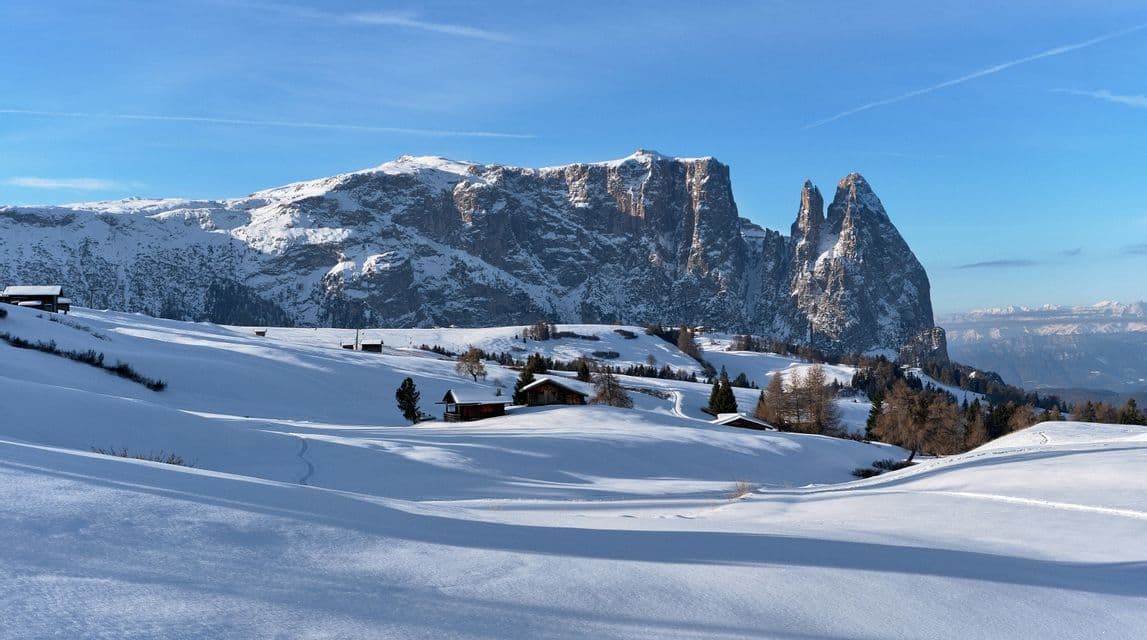 Un vasto paesaggio di colline innevate con baite di legno, sullo sfondo di una catena montuosa aspra sotto un cielo limpido.