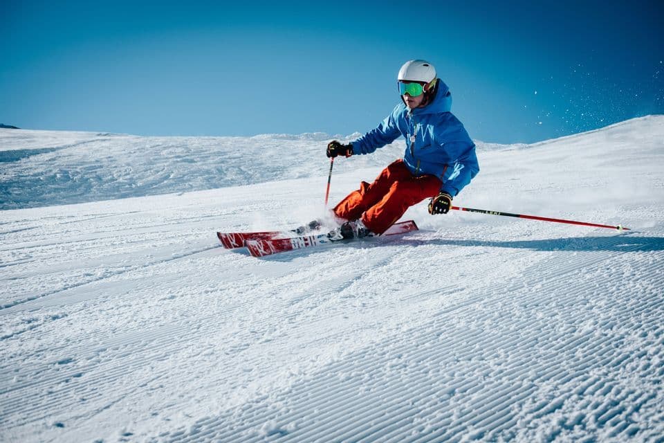 Uno sciatore con giacca e casco blu traccia una curva su una pista innevata battuta sotto un cielo azzurro limpido.