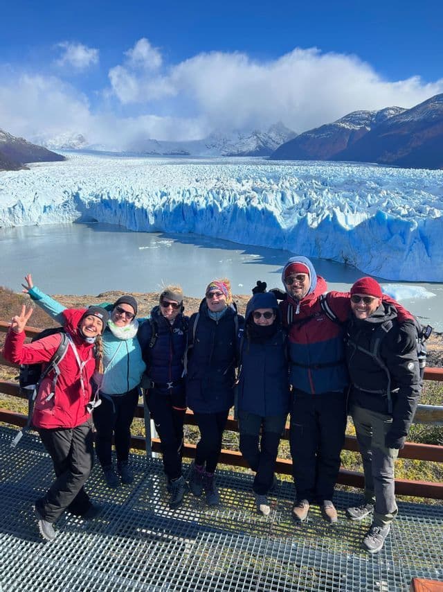 Eine WeRoad-Gruppe posiert für ein Foto auf einer Aussichtsplattform mit einem riesigen Gletscher und schneebedeckten Bergen im Hintergrund.