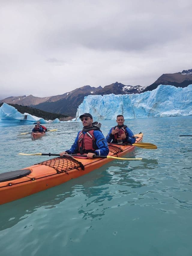 Eine WeRoad Gruppenreise beim Kajakfahren auf türkisfarbenem Wasser vor einem riesigen Gletscher und Bergen unter einem bewölkten Himmel.