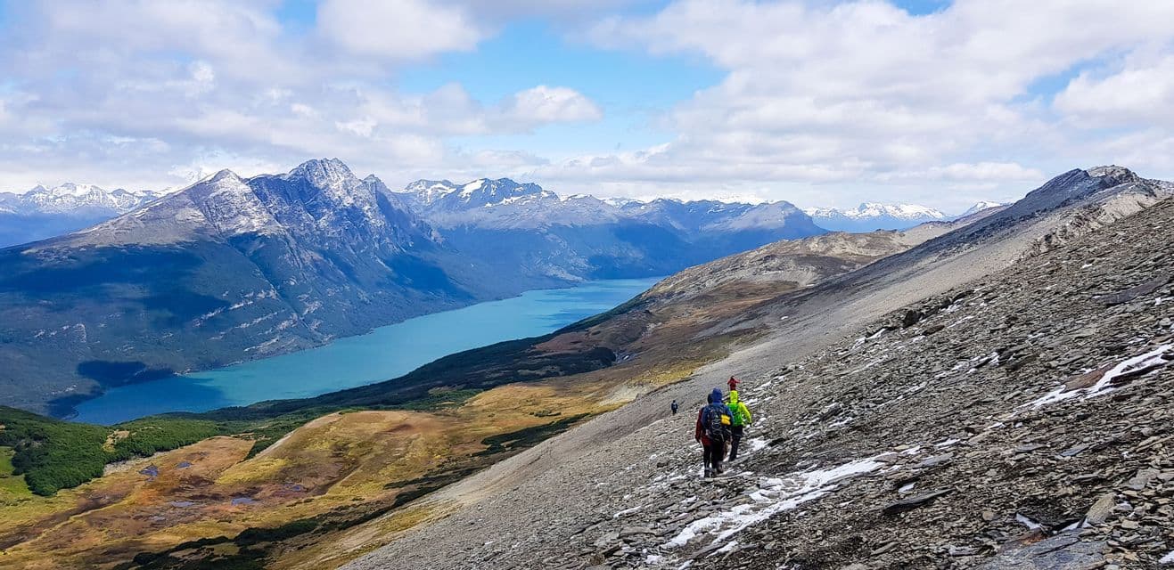 Eine WeRoad Gruppenreise wandert an einem steilen, felsigen Berghang mit Blick auf einen türkisfarbenen See und schneebedeckte Berge.