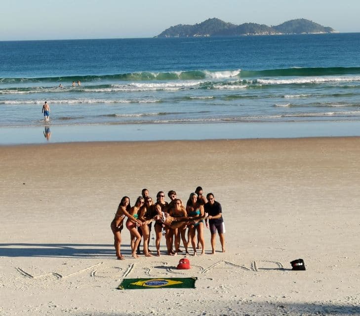 Eine WeRoad Gruppenreise posiert an einem Sandstrand, hält eine Person vor einer brasilianischen Flagge hoch, mit dem Ozean im Hintergrund.