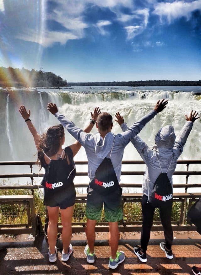 Drei Personen auf einer WeRoad-Gruppenreise, mit dem Rücken zur Kamera und erhobenen Armen vor einem großen, mächtigen Wasserfall.