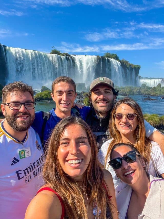 Eine WeRoad Gruppenreise von sechs Personen, die für ein Selfie vor einem großen Wasserfall unter klarem blauen Himmel lächeln.
