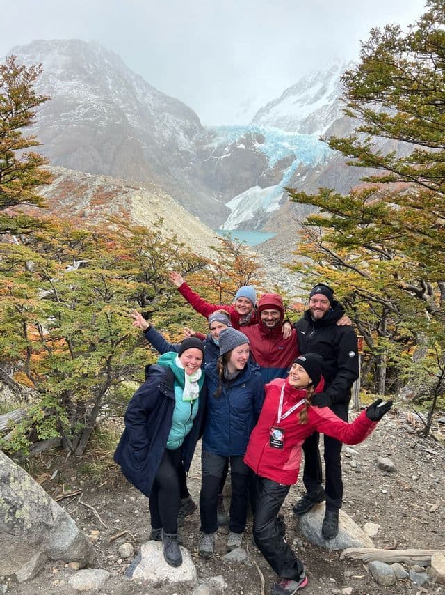 Eine WeRoad Gruppenreise lächelt für ein Foto auf einem Bergpfad, mit einem Gletscher und türkisen See im Hintergrund.