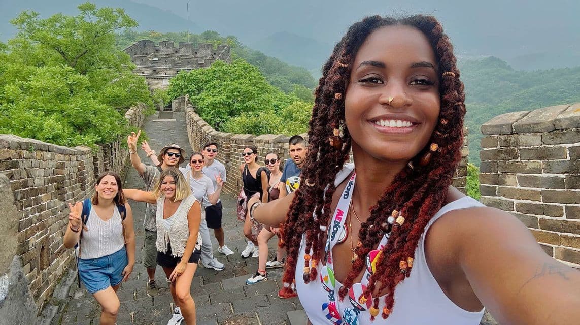 Eine Frau mit Zöpfen macht ein lächelndes Selfie mit ihrer WeRoad-Reisegruppe auf einer großen Steinmauer, die sich durch grüne, neblige Hügel schlängelt.