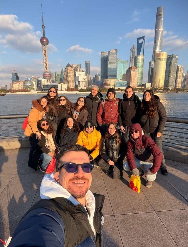 Ein Mann macht ein Selfie mit seiner WeRoad-Gruppe, während sie gemeinsam an einer Uferpromenade vor einer modernen Stadtsilhouette posieren.