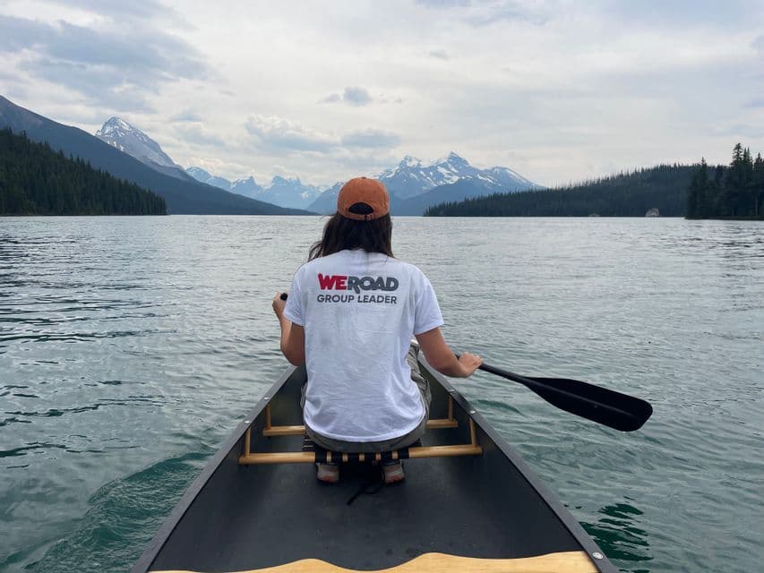 Un Capogruppo WeRoad visto da dietro, che pagaia in canoa su un lago verso distanti montagne innevate.
