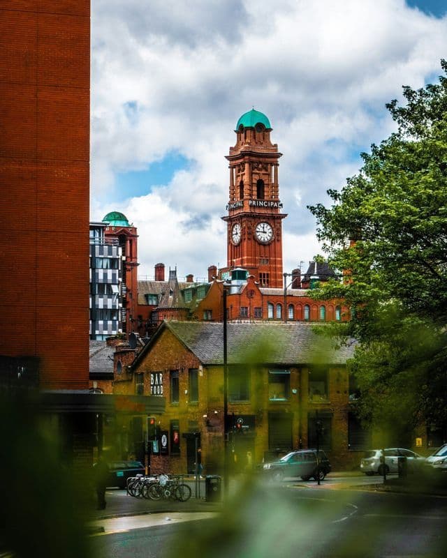 Una alta torre dell'orologio in mattoni rossi con una cupola verde si erge sopra gli edifici della città e il traffico stradale sotto un cielo nuvoloso.