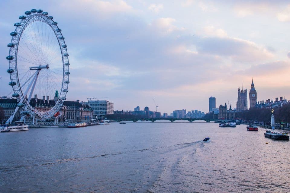 Una grande ruota panoramica si erge sulla riva di un fiume con barche che navigano accanto agli edifici della città al crepuscolo